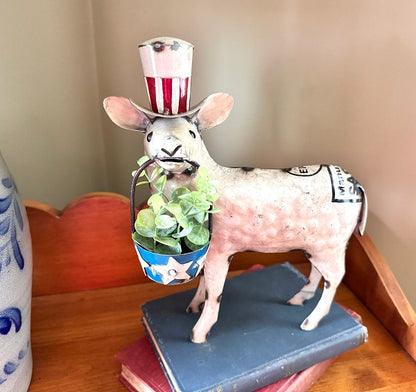 Decorative sheep figurine wearing a top hat with a flag design, holding a plant, on a wooden surface.