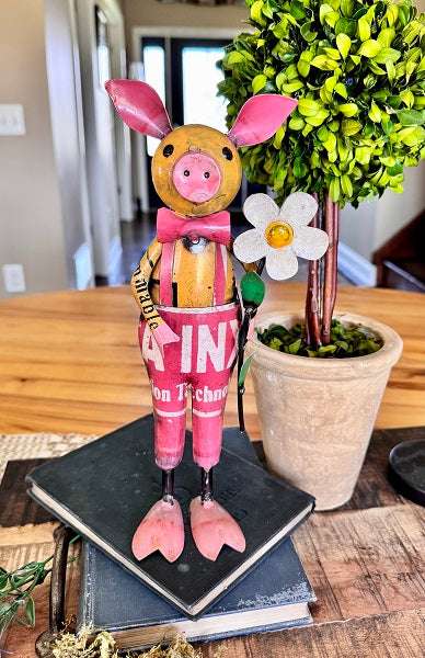 Decorative pig figurine with pink outfit on a wooden table with a plant in the background.