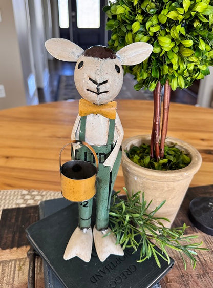 Decorative figurine of a sheep in overalls holding a bucket, placed on a table with a plant in the background.