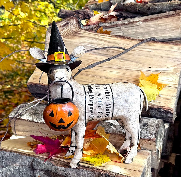 Decorative sheep statue with a witch hat and pumpkin, surrounded by autumn leaves and stacked firewood.