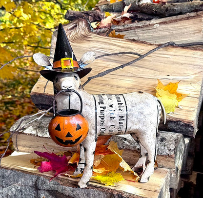 Decorative sheep statue with a witch hat and pumpkin, surrounded by autumn leaves and stacked firewood.