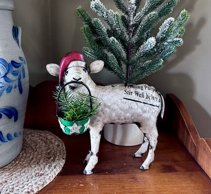 Decorative sheep figurine with a Santa hat and green basket, standing on a wooden surface next to a blue and white vase and a small Christmas tree.