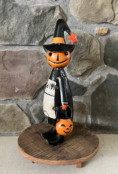 Halloween-themed figurine with a pumpkin face and witch hat on a wooden base against a stone wall.