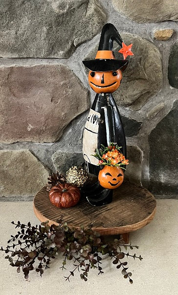 Halloween-themed  figurine with a jack-o'-lantern face, black hat, and pumpkin pail on a wooden stand against a stone wall.