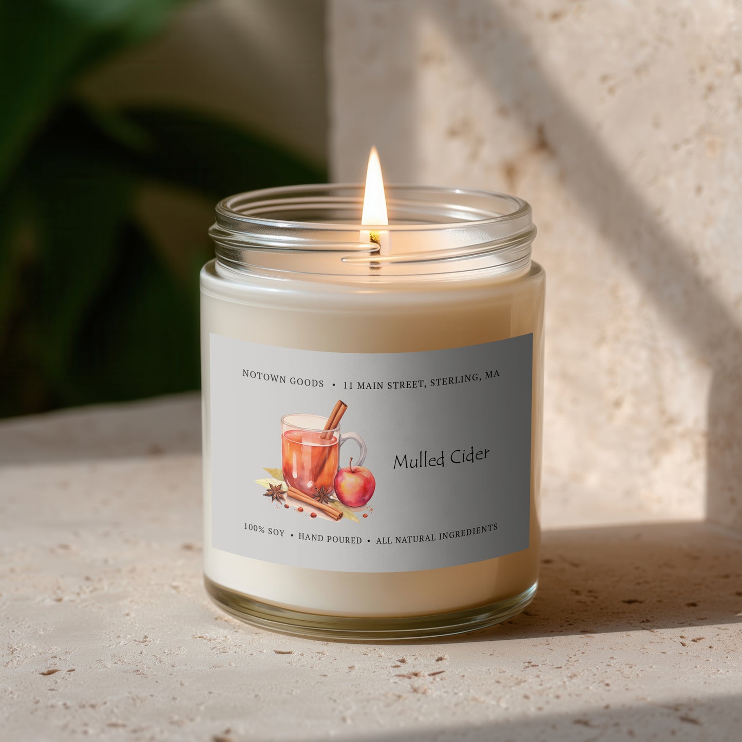 Clear jar candle with white wax and a decorative label on a blurred outdoor background.