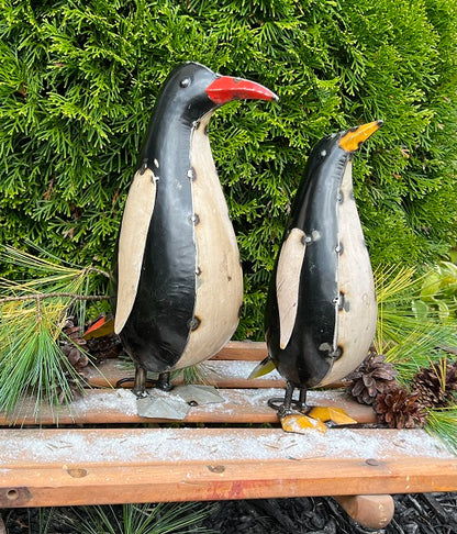 Two penguin figurines on a wooden platform with a green bush background
