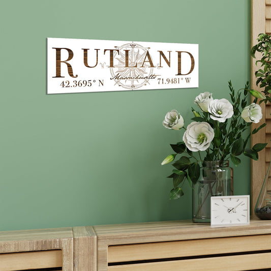 Decorative sign with 'Rutland, Massachusetts' and coordinates on a green wall above a wooden surface with flowers and a clock.