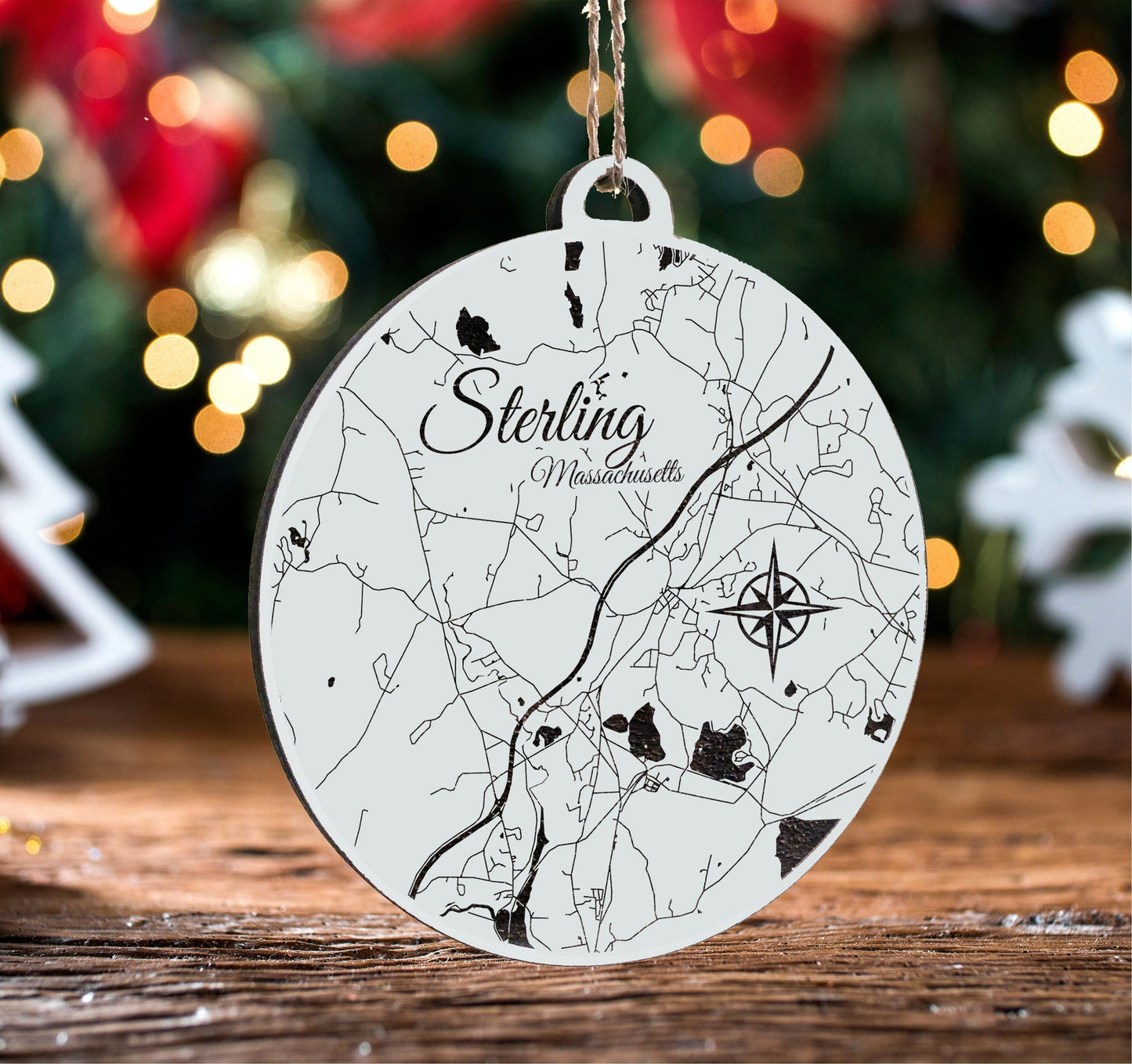 Map ornament with 'Sterling, Massachusetts' text on a wooden surface with Christmas lights in the background