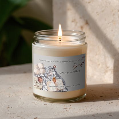 Clear jar candle with white wax and a decorative label on a blurred outdoor background.