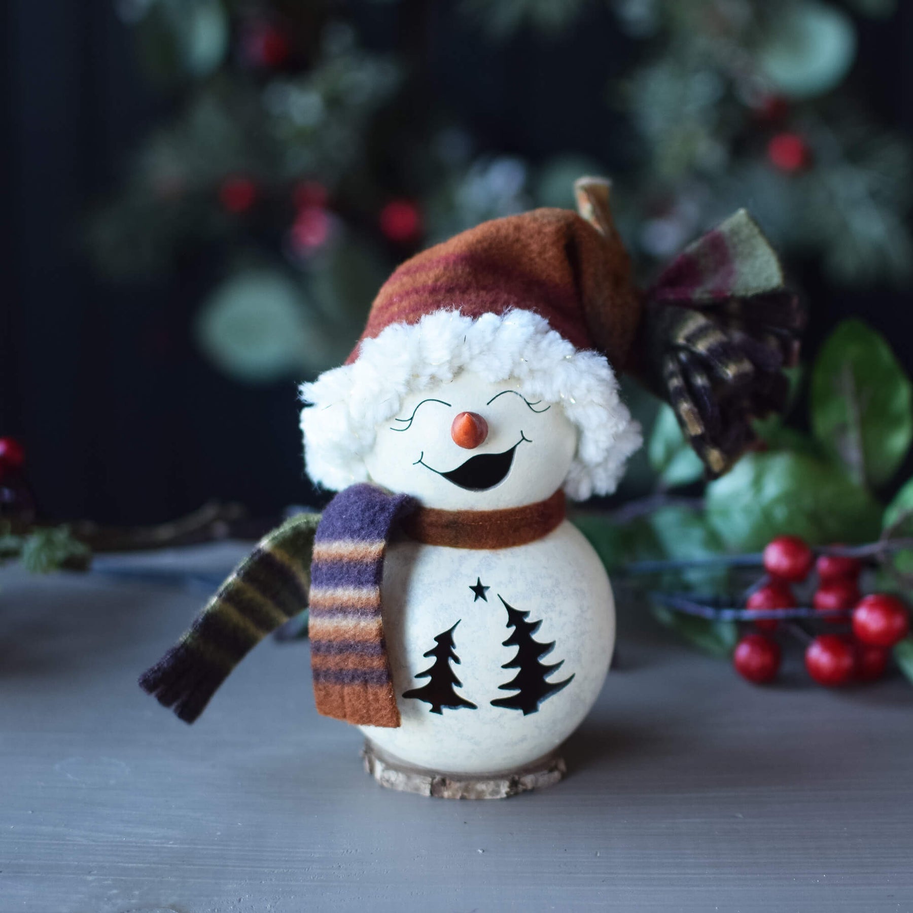 Decorative snowman figurine with a scarf and hat on a table with a blurred Christmas tree background.
