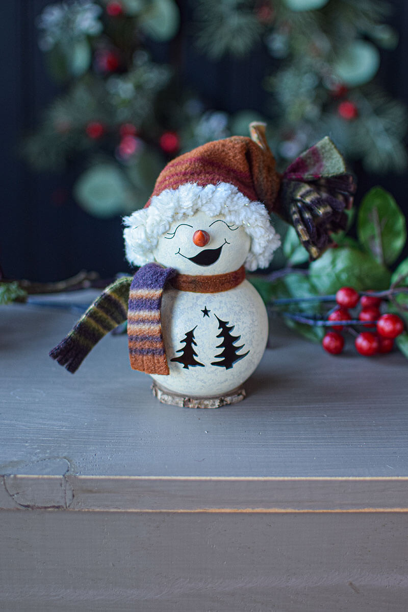 Decorative snowman figurine with a colorful scarf and hat on a wooden surface with a Christmas tree in the background.