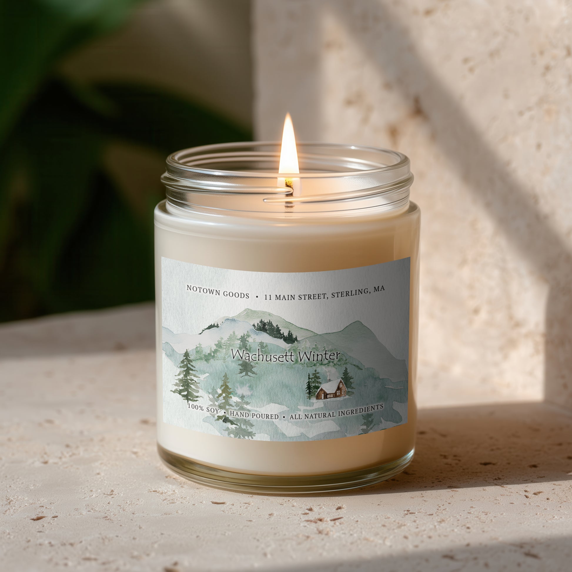 Clear jar candle with white wax and a decorative label on a blurred outdoor background.