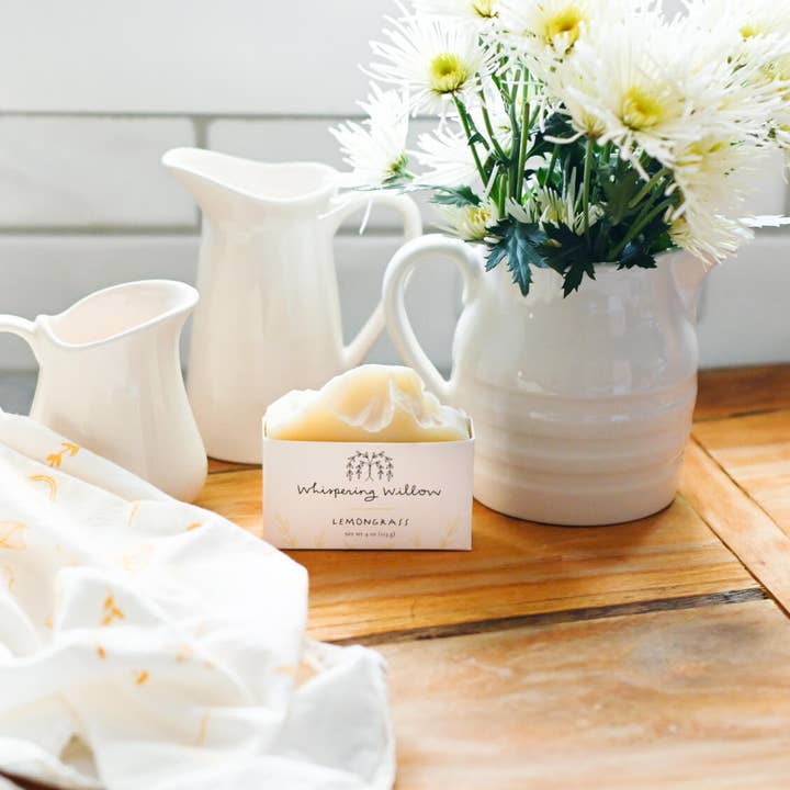 Soap bar with packaging on a wooden surface next to white ceramic pitchers and flowers.