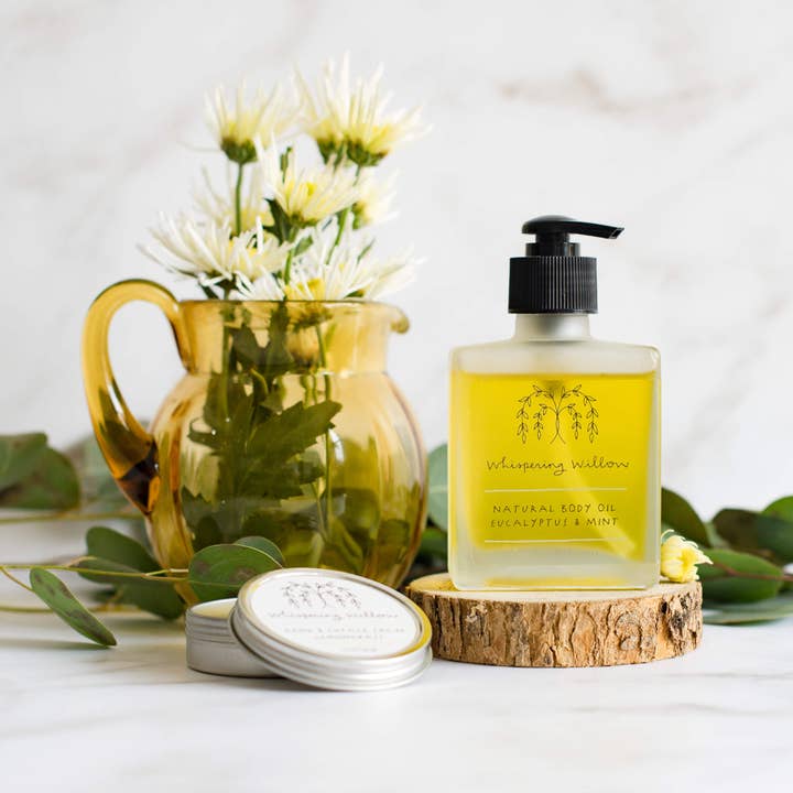 Bottle of natural body oil on a wooden coaster with a pitcher of flowers and leaves in the background.