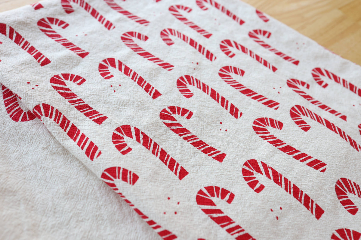 A close-up of a white fabric tea towel, printed with a repeating red candy cane pattern on a light wood surface.