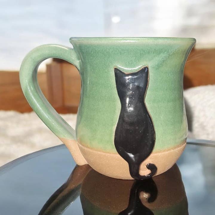 Green ceramic mug with a black cat design on a reflective surface