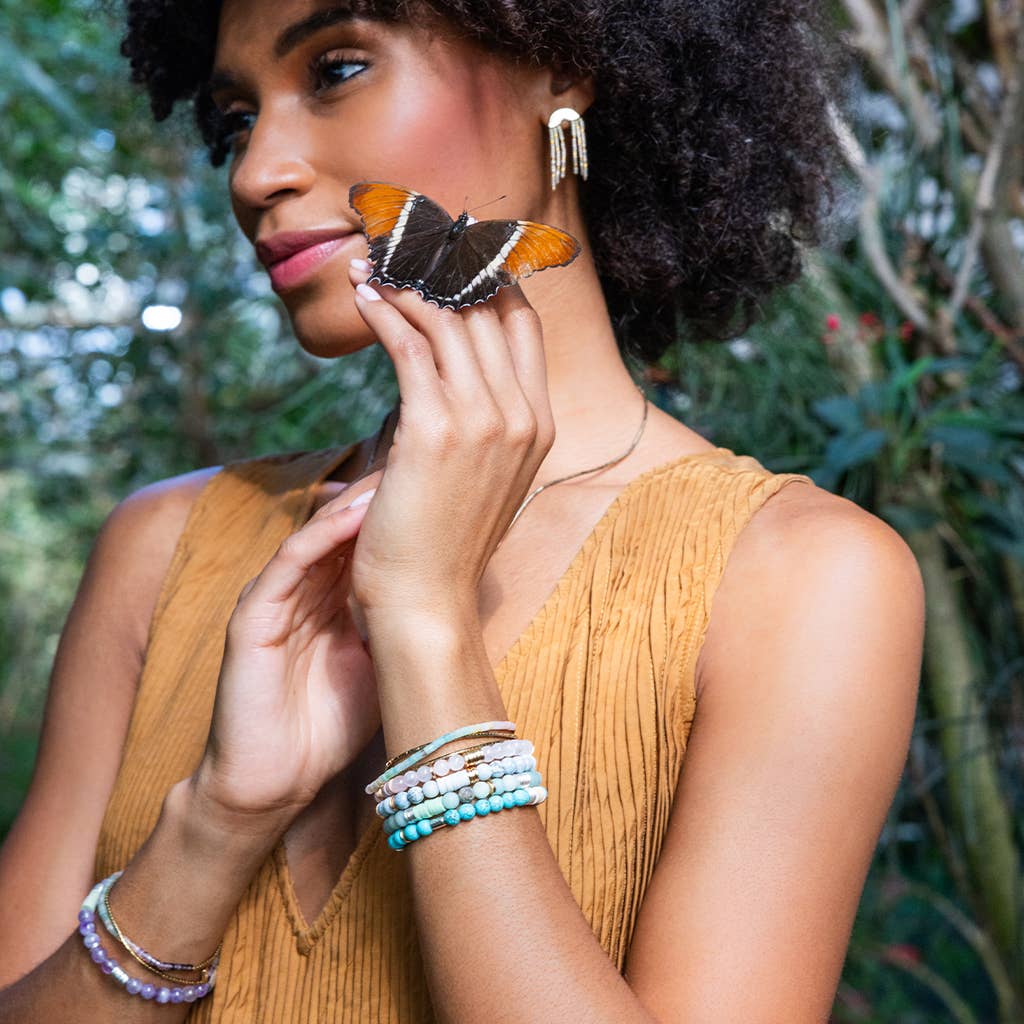 Woman with a butterfly on her finger, wearing a mustard yellow top and multiple bracelets, with a blurred natural background.