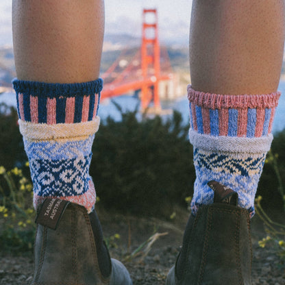 A close-up of a person's legs wearing colorful knit socks with a Fair Isle pattern in pink, blue, cream, and navy, paired with brown Chelsea boots, with a blurred Golden Gate Bridge in the background.