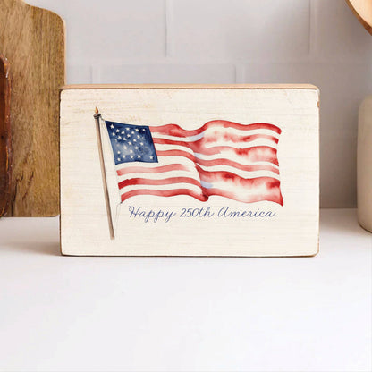Wooden block with American flag design and 'Happy 250th America' text on a white surface.
