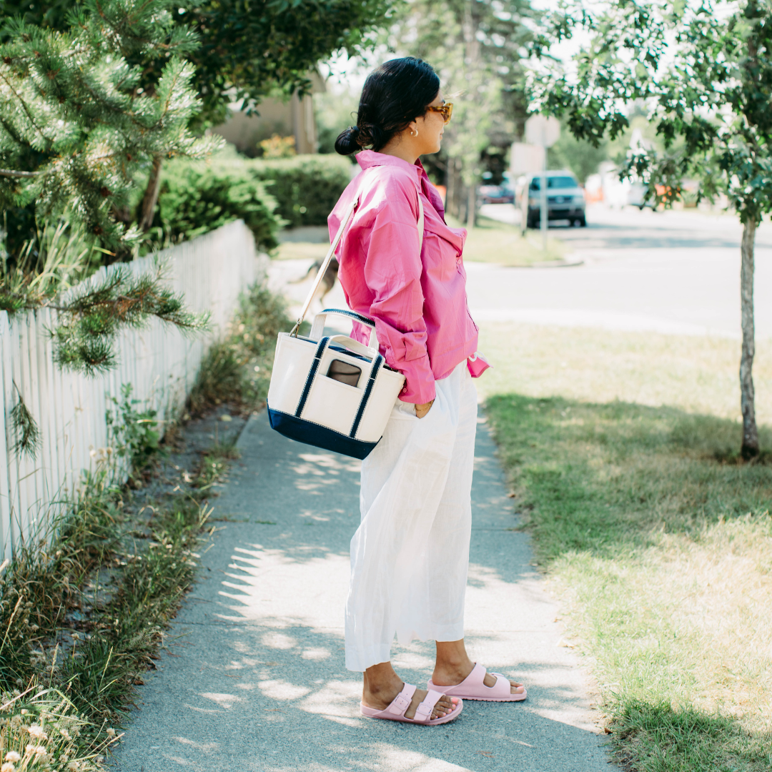 Woman in pink jacket and white pants walking on a sidewalk with a tote bag.