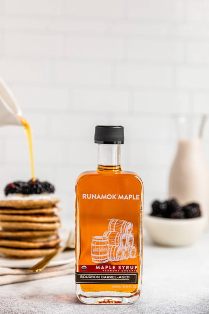 A bottle of Runamok Maple Bourbon Barrel-Aged maple syrup stands on a countertop in a kitchen setting, with pancakes topped with syrup and berries blurred in the background.