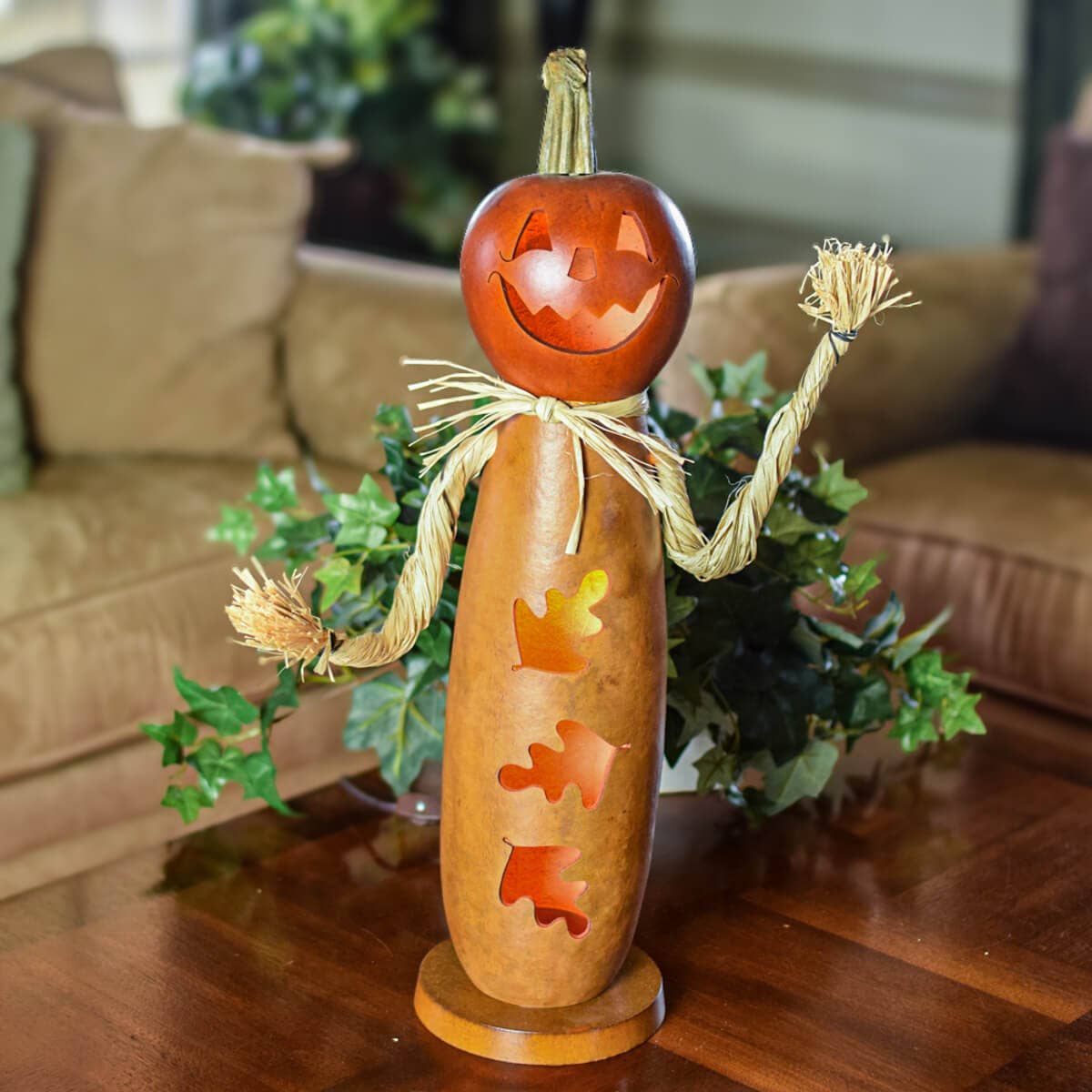 A Halloween decorative gourd figure standing on a round wooden base, with a carved orange pumpkin head, straw-colored rope arms and hair, and cutout details on the body, photographed on a wooden table with a couch and greenery in the background.