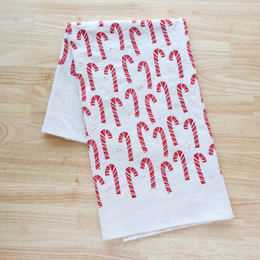 A white fabric tea towel with a repeating red candy cane pattern, folded on a light wood grain surface.