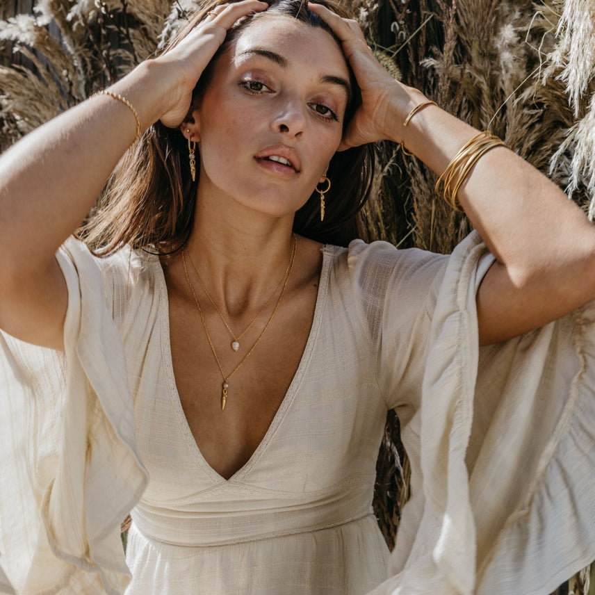 Woman in a flowing white dress with gold jewelry against a natural background