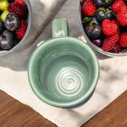 Green ceramic mug on a wooden surface with bowls of berries in the background