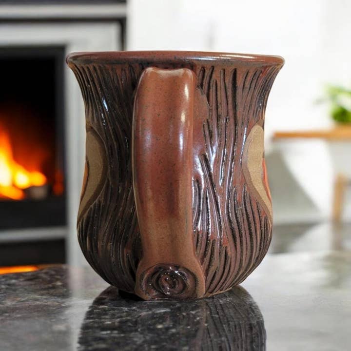 Brown ceramic mug with decorative handle on a reflective surface, fireplace in the background