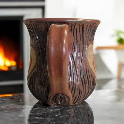 Brown ceramic mug with decorative handle on a reflective surface, fireplace in the background