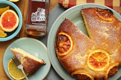 A slice of orange-glazed cake on a light blue plate, with a bottle of Runamok maple syrup nearby and additional orange slices on a wooden table.