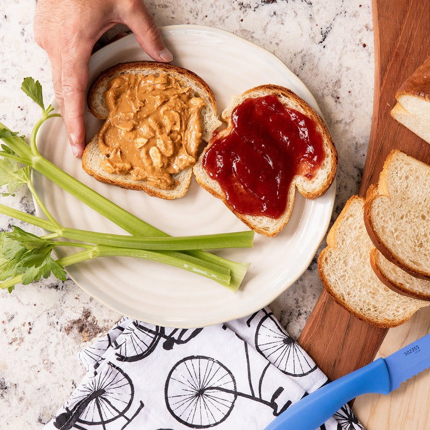 Peanut butter and strawberry jam sandwich on a plate with celery sticks, surrounded by bread slices and a knife.