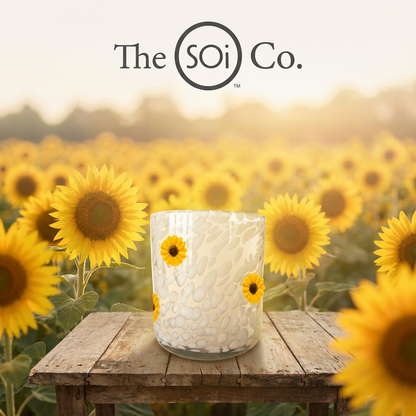 Glass candle holder with sunflower design on a wooden table in front of a sunflower field, featuring 'The SOi Co.'