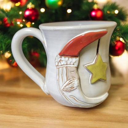 Decorative mug with a moon and star design on a wooden surface with a Christmas tree in the background.