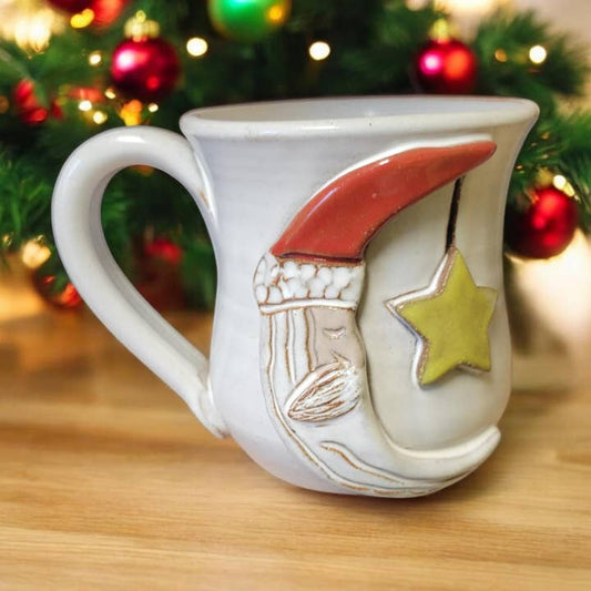 Decorative mug with a moon and star design on a wooden surface with a Christmas tree in the background.