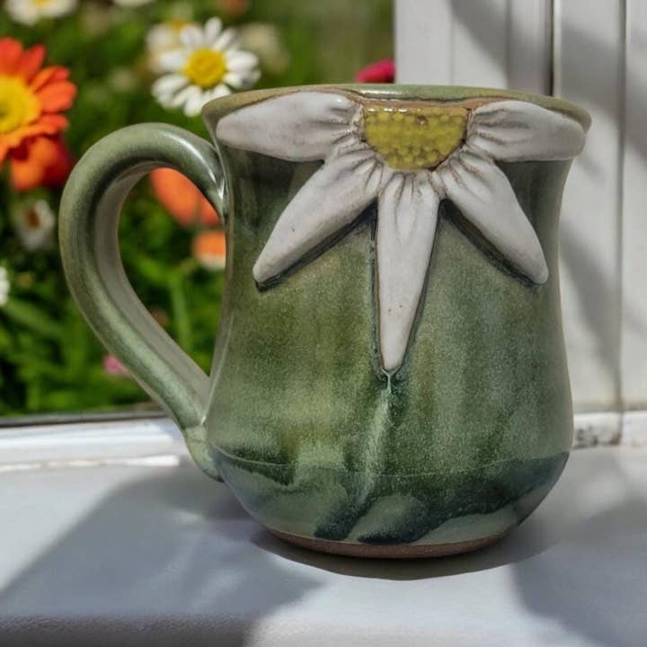 Green ceramic mug with a floral design on a windowsill with flowers in the background