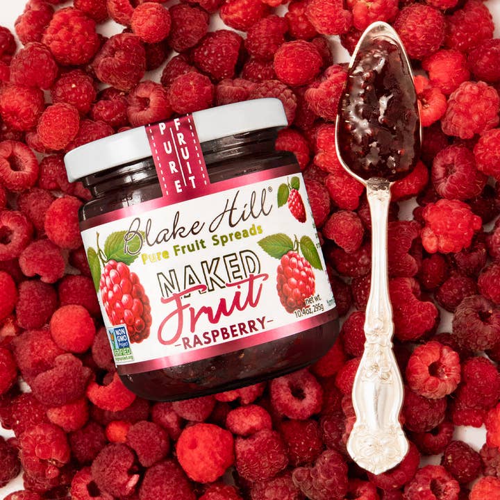 Jar of Blake Hill Naked Fruit Raspberry spread surrounded by raspberries with a spoonful of spread.