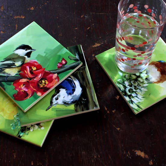 Set of ceramic coasters with bird and floral designs on a dark wooden surface with a glass of water. 