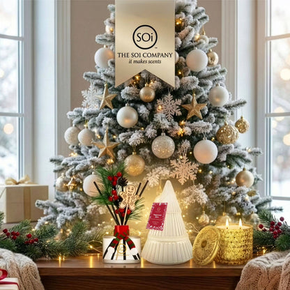 A decorated Christmas tree in a cozy indoor setting, with white and gold ornaments, warm string lights, snowflake and star ornaments, and a hanging banner reading 'The Soi Company'. In front of the tree on a wooden table are holiday decor items including a white tree-shaped candle holder, a gold textured candle, a small gift box with red ribbon, and green garlands.