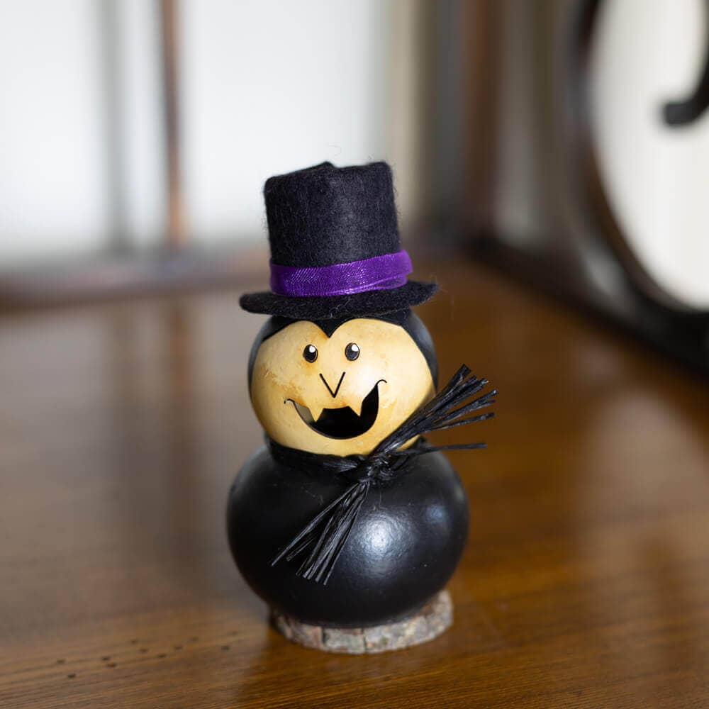 A small Halloween decorative figure shaped like a gourd, with a black painted body, a beige face, a black felt top hat with a purple band, and a small black twig staff, standing on a wooden surface.