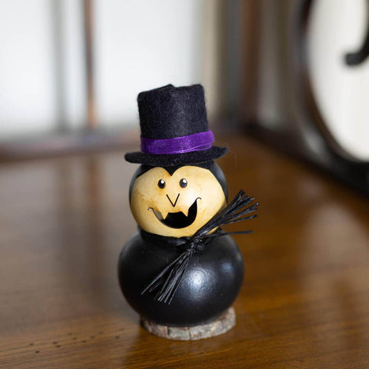 A small Halloween decorative figure shaped like a gourd, with a black painted body, a beige face, a black felt top hat with a purple band, and a small black twig staff, standing on a wooden surface.