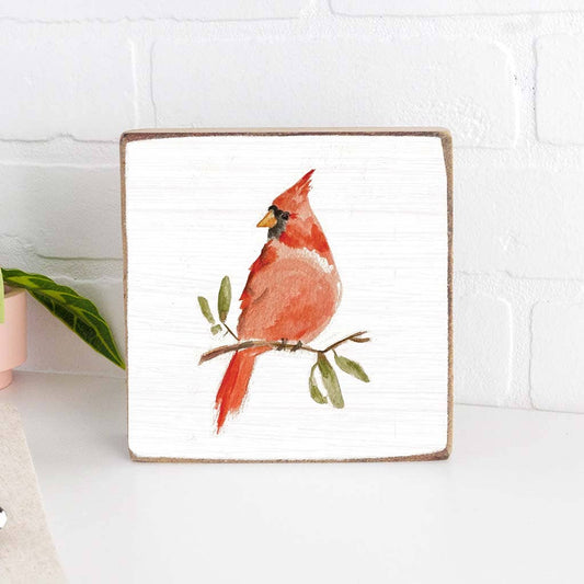 Wooden block with a red cardinal bird on a branch with leaves against a white brick wall.