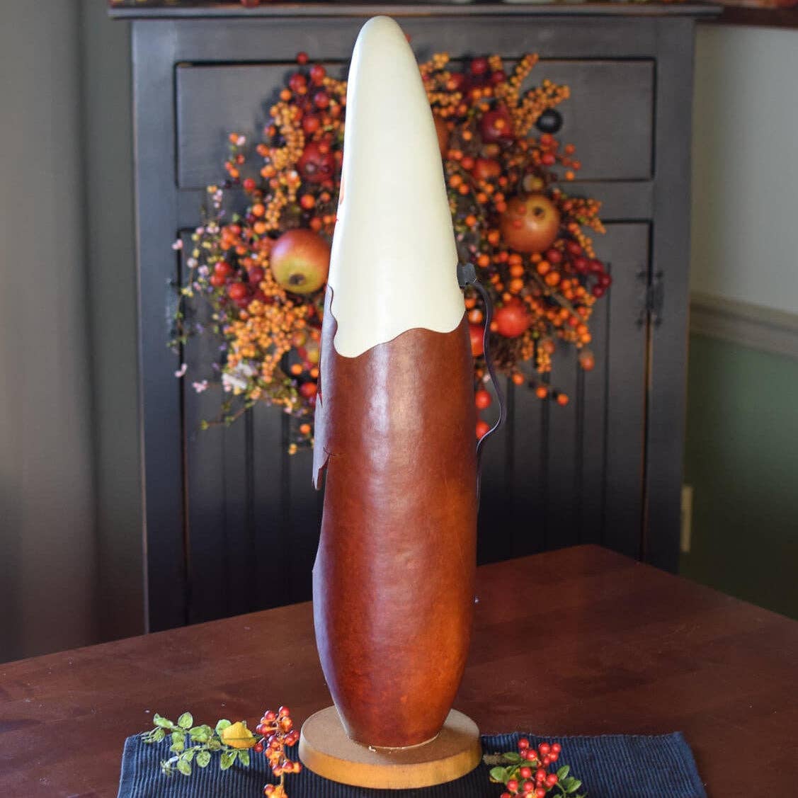 A tall, sculptural decorative gourd with a glossy reddish-brown lower section and a smooth off-white upper section, displayed on a wooden base on a dark wood table with a fall-themed wreath in the background.