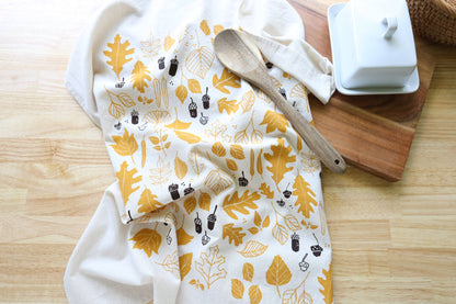 A white kitchen towel with a mustard yellow and dark brown autumn leaf pattern, styled on a light wood surface next to a wooden spoon and a white butter dish.