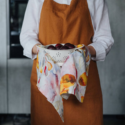 Person wearing a brown apron holding a colander with plums and a colorful towel.
