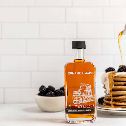 A bottle of Runamok Maple bourbon barrel-aged syrup stands on a white countertop in a kitchen setting, with a plate of pancakes dripping syrup and a bowl of blackberries blurred in the background.