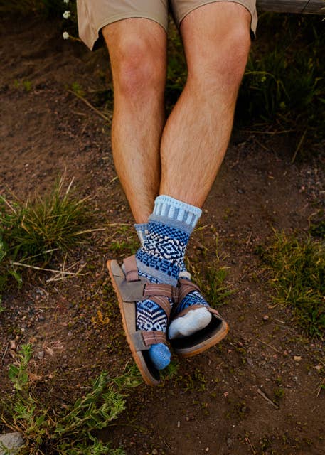 A close-up lifestyle shot of a person's legs crossed, wearing patterned blue and white crew socks, brown leather sandals, and khaki shorts while sitting outdoors on a dirt path.