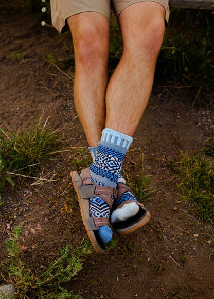 A close-up lifestyle shot of a person's legs crossed, wearing patterned blue and white crew socks, brown leather sandals, and khaki shorts while sitting outdoors on a dirt path.