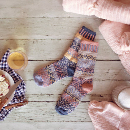 A pair of patterned knit socks laid flat on a light wood surface, surrounded by soft pink textiles and small breakfast props. The socks have a multicolor Fair Isle style design with dusty pink, navy, beige, and mustard tones.